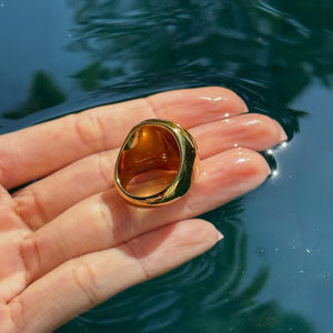 A gold signet-style ring with a polished amber-brown oval face, held on a person's fingers over dark rippling water.
