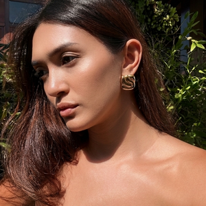 A woman outdoors wearing a chunky gold geometric hoop earring, with lush green foliage and a house in the background.